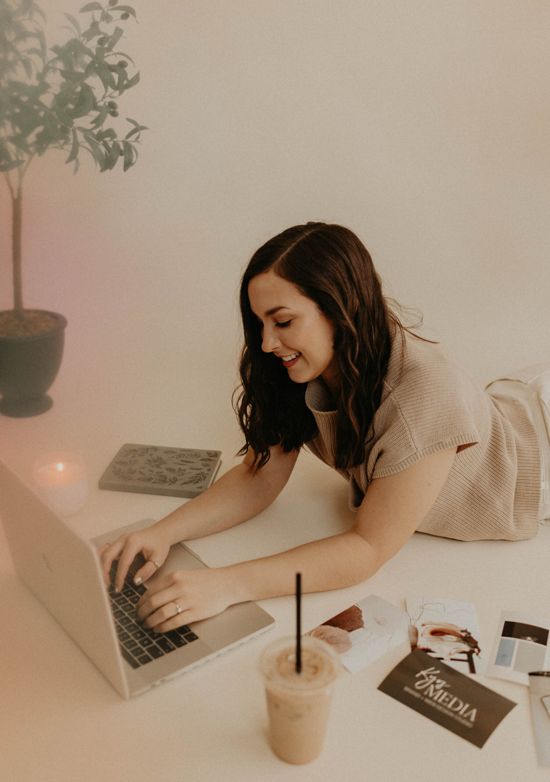 Brunette woman lying on the floor surrounded by her laptop, an iced coffee, a green notebook, a lit candle, and photos.