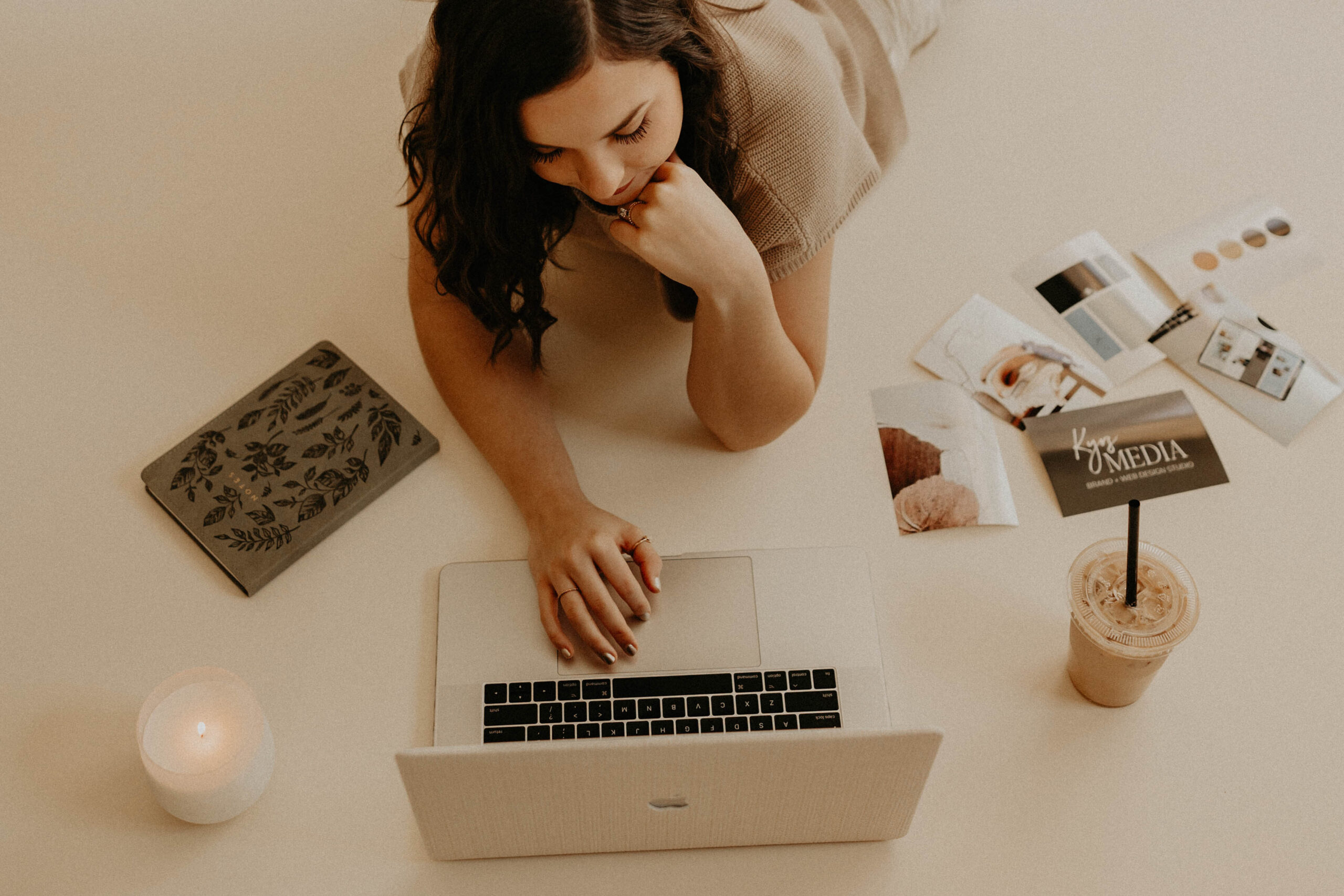 Brunette woman lying on the floor surrounded by her laptop, an iced coffee, a green notebook, a lit candle, and photos.